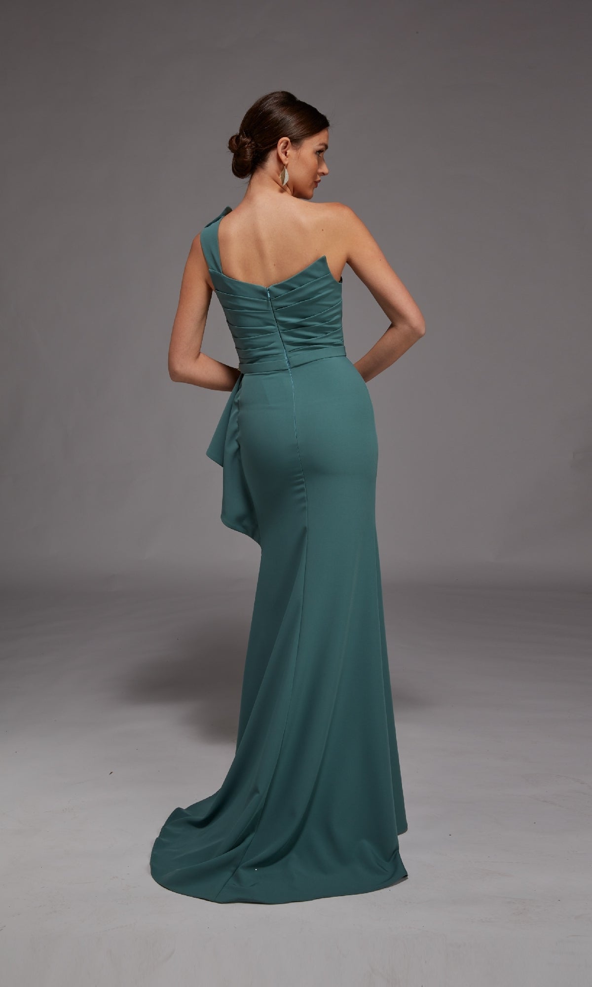 A woman in the Alyce 27812 long formal dress, featuring a teal hue, one-shoulder neckline, and crystal-beaded accent, stands with her back to the camera against a plain gray background.